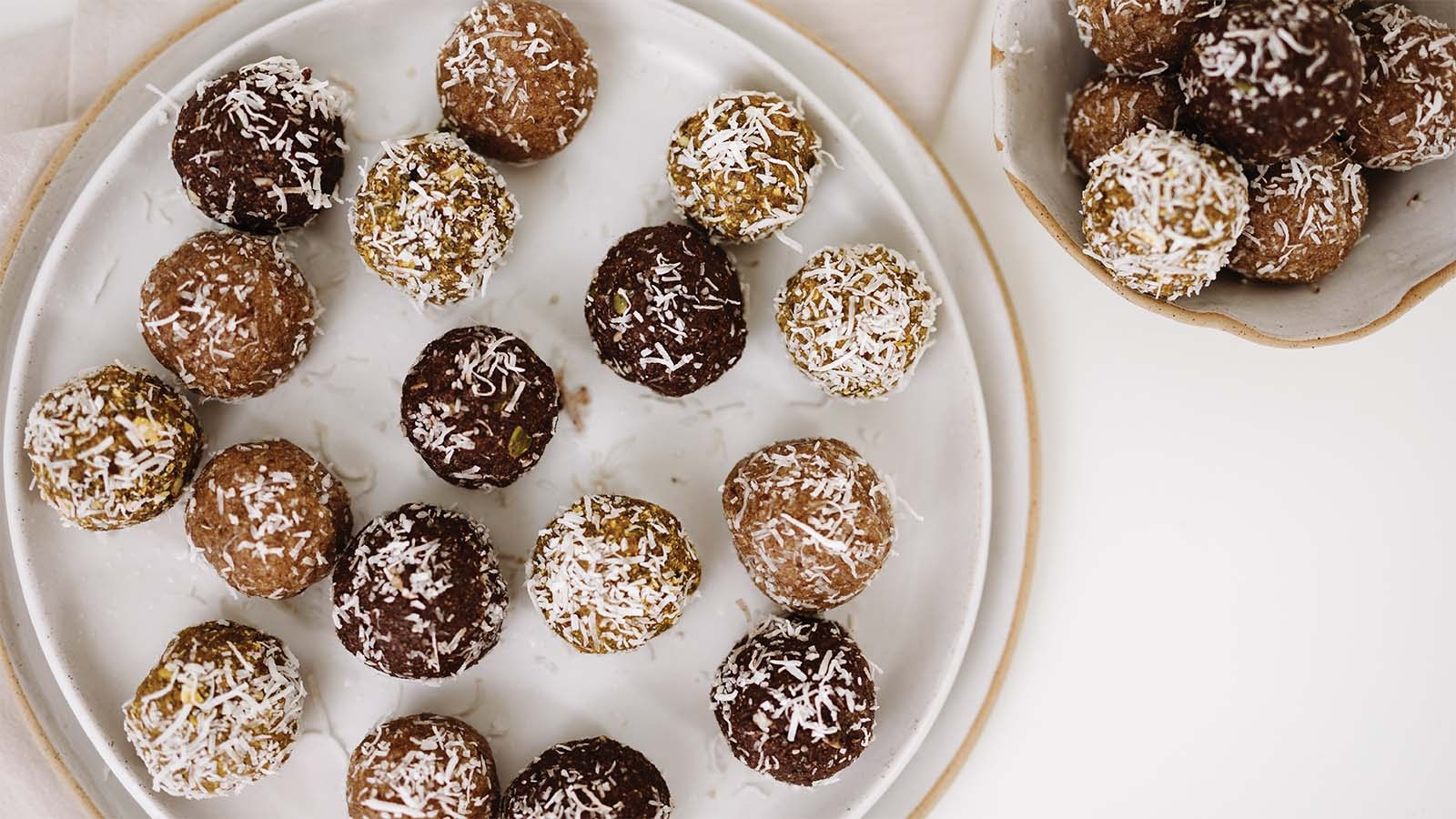 Coconut-coated chocolate, tan, and pistachio energy balls arranged on white plates.