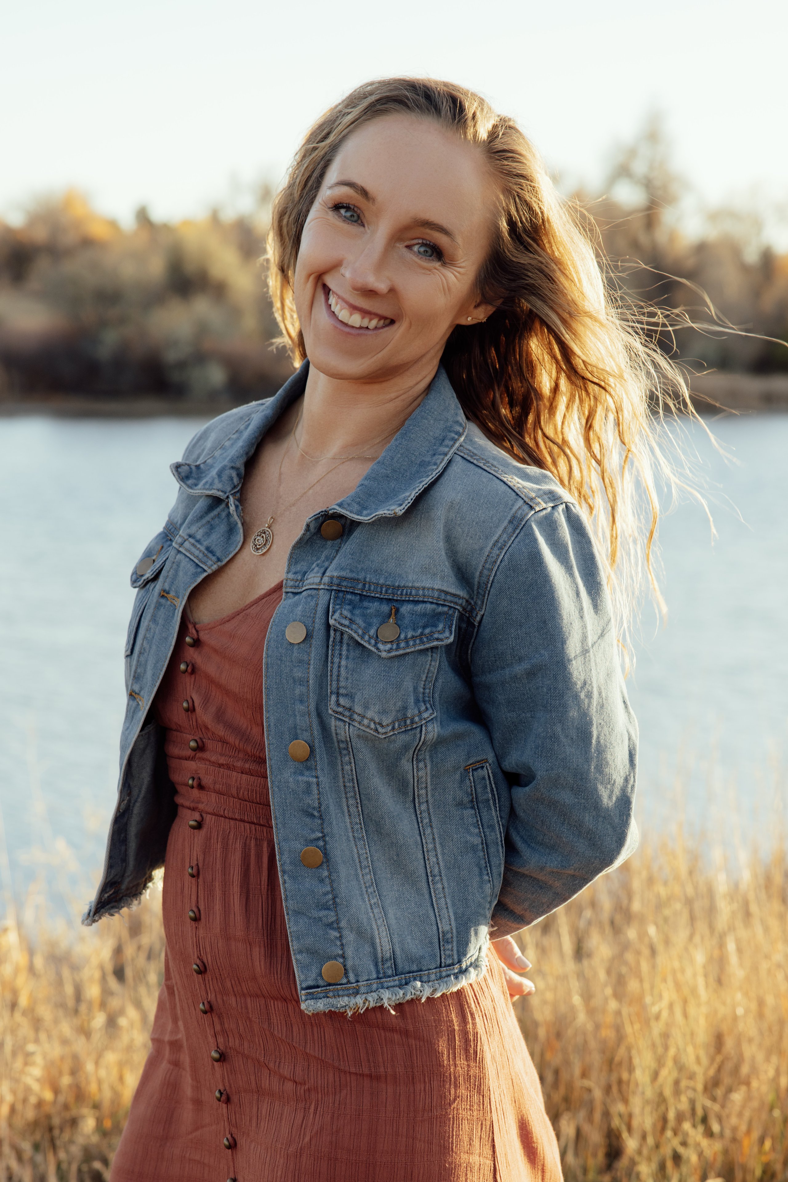 A smiling woman with windblown wavy hair wears a denim jacket and rust dress by a lake in golden sunlight.